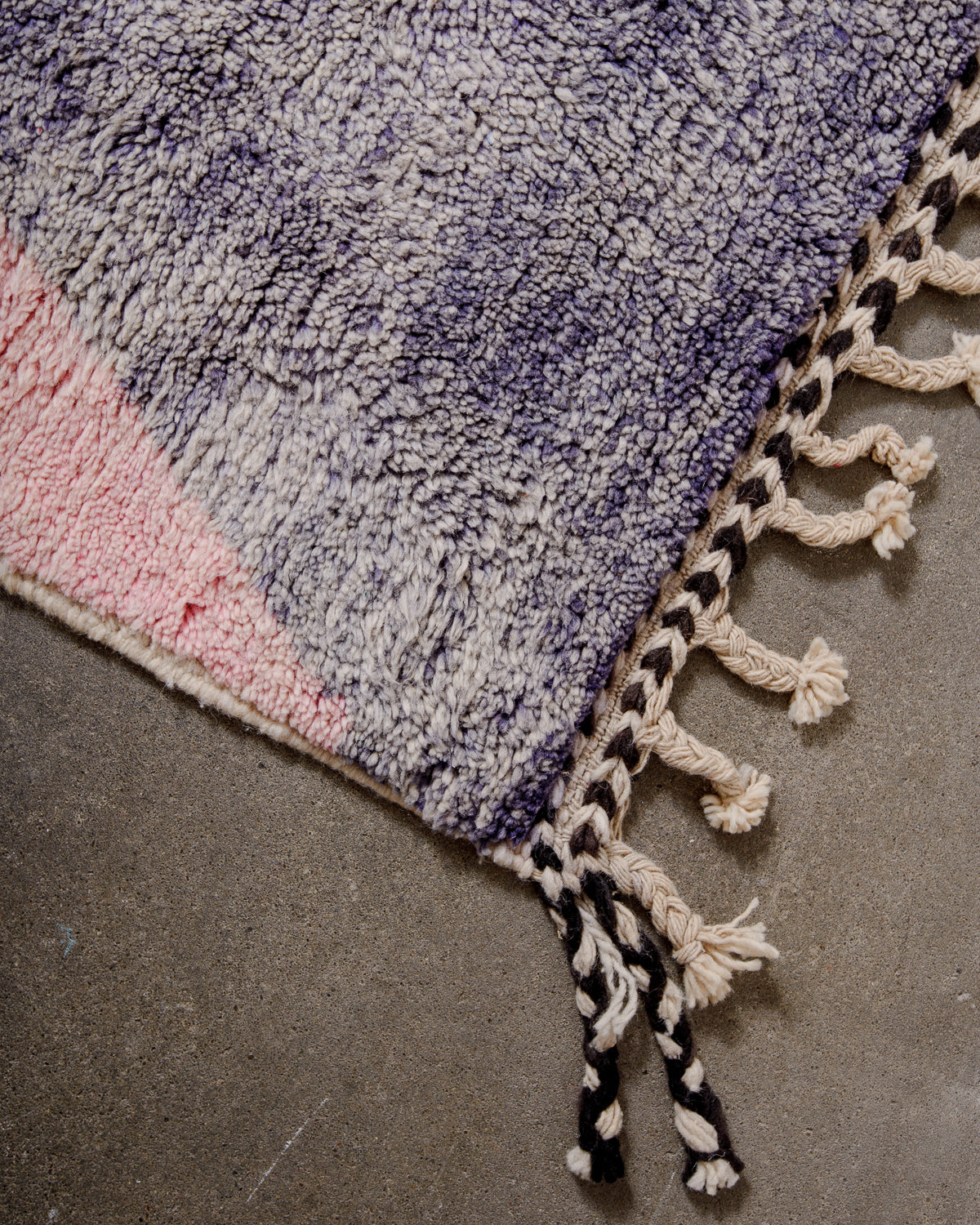 Corner of Berber rug made of lavender and pink wool with black and white details on the edge and natural wool tassels.